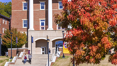 a bright red and orange tree in the foreground with one of the academic buildings
                        and students walking to class in the background