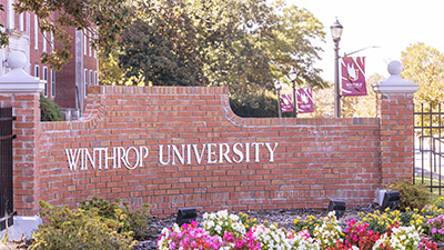 a brick Winthrop University sign surrounded by brightly colored flowers on a sunny
                        day