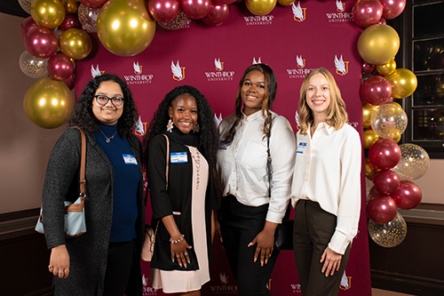 Four students in professional attire standing in front of the Winthrop logo 