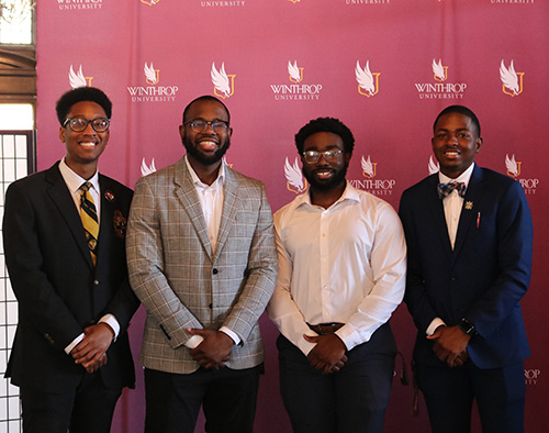 Four students in professional attire posing in front of the Winthrop logo