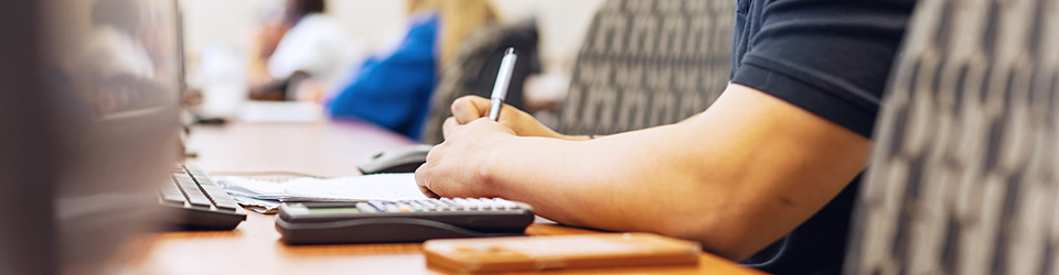 a student writing calculations on a notebook page with a calculator beside them