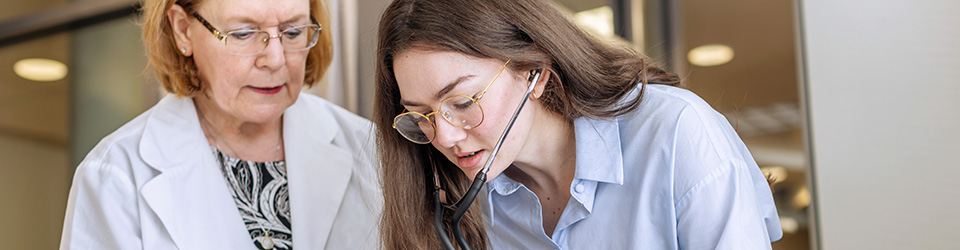 a professor in a lab coat working with a student using a stethoscope