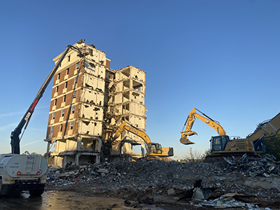 Blue sky with cranes tearing into tall high-rise brick building. Rubble scattered
                        all around