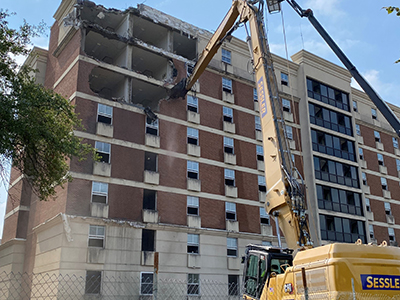 Yellow crane tearing away at tall brick dorm building