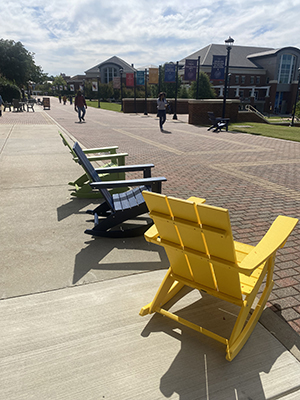 Green, blue, and yellow Adirondack chairs on brick walkway, blue sky background