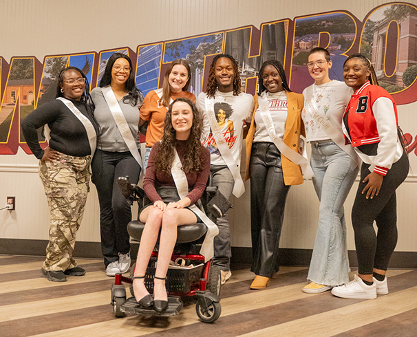 a group of students including one in a wheelchair gathered in front of a Winthrop
                              mural