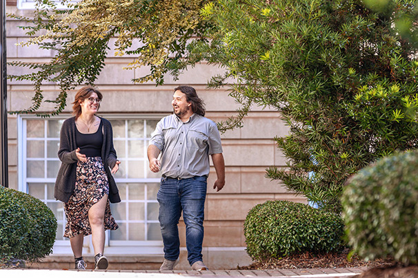 two students walking and laughing together on campus beside green trees and shrubs