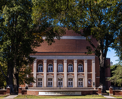 a photograph of the Winthrop University sign with Byrnes Auditorium in the background. It is a sunny spring day and there are flowers blooming beside the sign