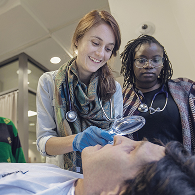 Two women wearing stethoscopes and gloves looking at a patient's eye