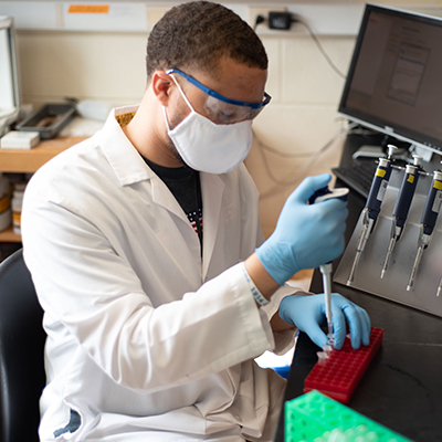 A man in a lab coat and protective gear pipetting something into tubes