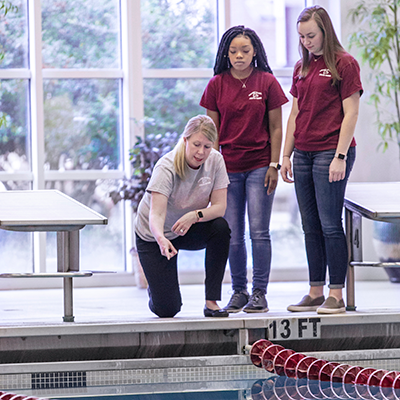 Three women standing at the side of a pool with one woman pointing toward the line
                           floats and speaking