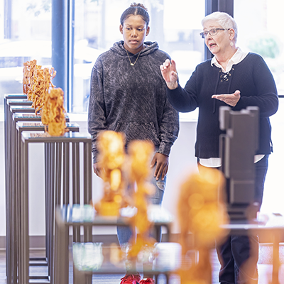 Two women talking in a gallery surrounded by small gold statues on pedestals