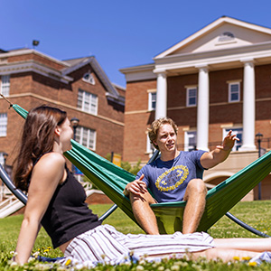 three students sitting on the grass. one is playing guitar and another is singing