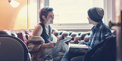 Two students talking while sitting on a leather couch in a cozy room with bookshelves.